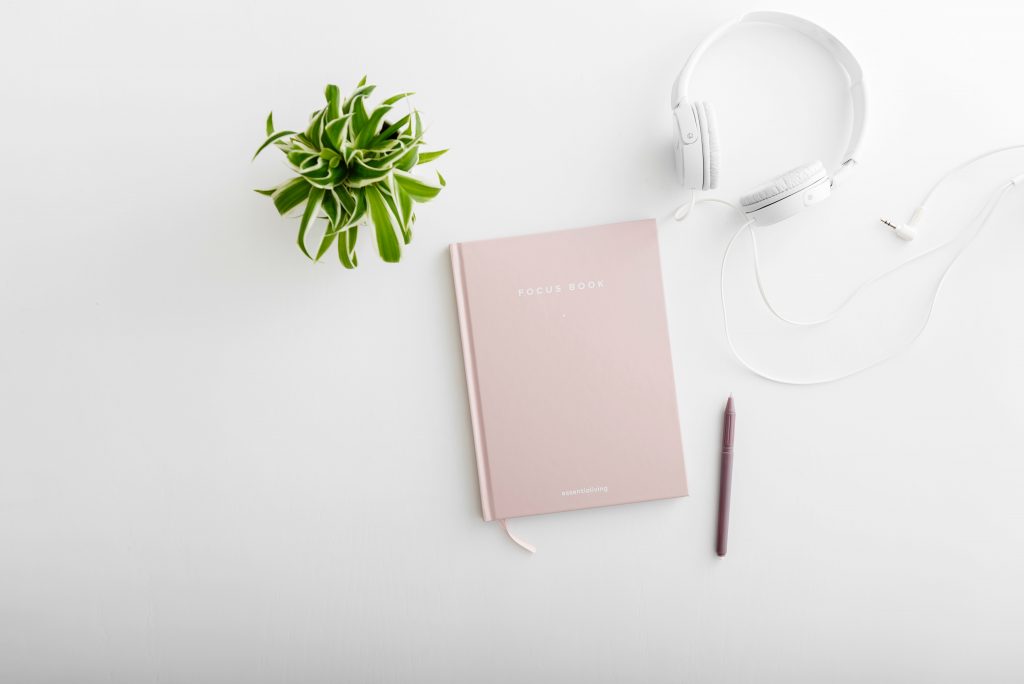 A diary on a white table top next to a pen, a potted plant, and headphones.