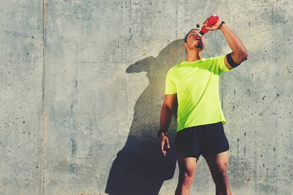 Young tired athlete refreshing with energy drink while standing against cement wall background