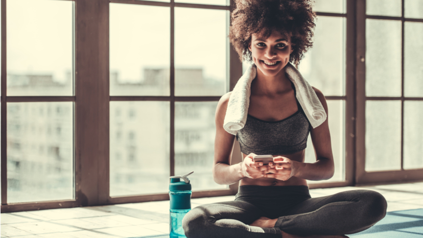 Woman in sportswear is using a smart phone, looking at camera and smiling while sitting on yoga mat.