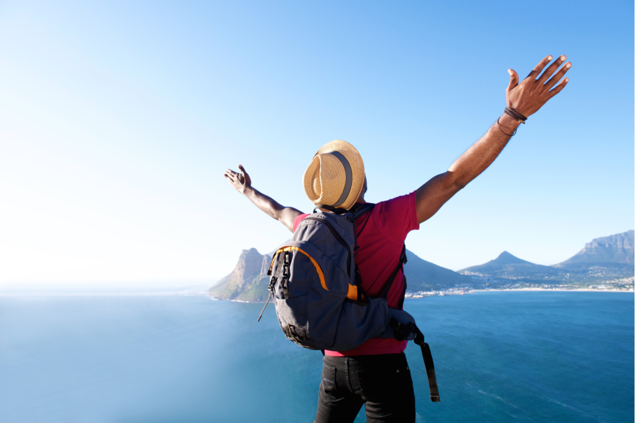 Rear view portrait of a guy with hat and backpack standing outdoors with arms spread open enjoying ecotherapy.