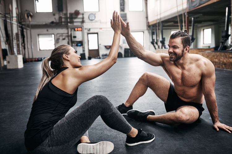 Two fit young people in sportswear smiling and high fiving together while sitting on a gym floor after working out
