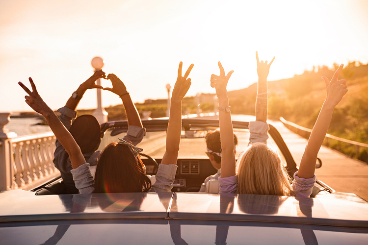 A group of happy people/friends while driving on the road with their hands up facing the sunset.