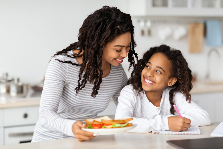 Cute black mother and daughter eating sandwiches at kitchen.