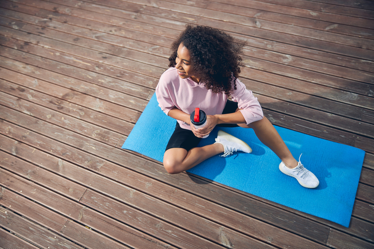 High angle photo of a woman sitting on yoga mat and holding bottle of water.