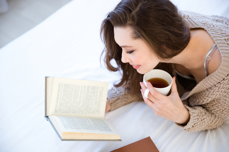 Woman reading book and drinking coffee on bed.