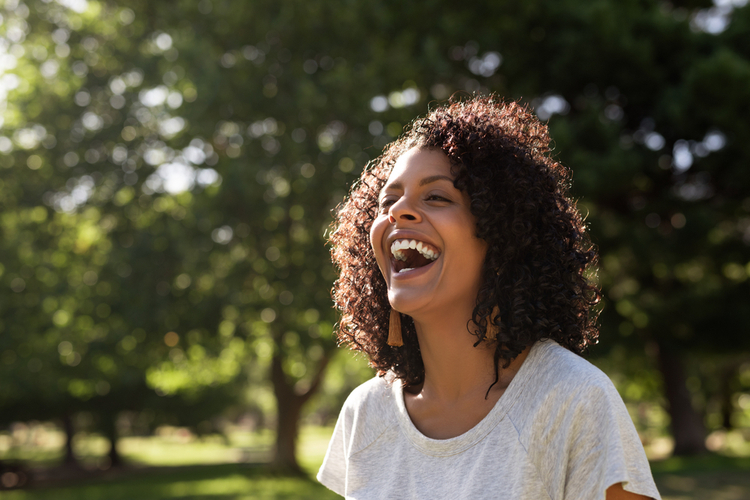 Woman with curly hair laughing while standing outside in a park knowing how to live a happy life.