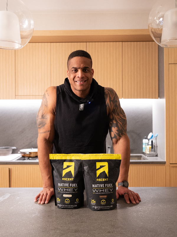 Corey posing with two bags of Ascent protein in the kitchen